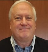 Older man with white hair wearing a black blazer and blue checkered shirt against a brown background